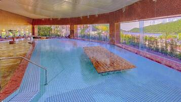 An indoor swimming pool with a wooden deck, tiled floor, and large windows offering a view of greenery and buildings.