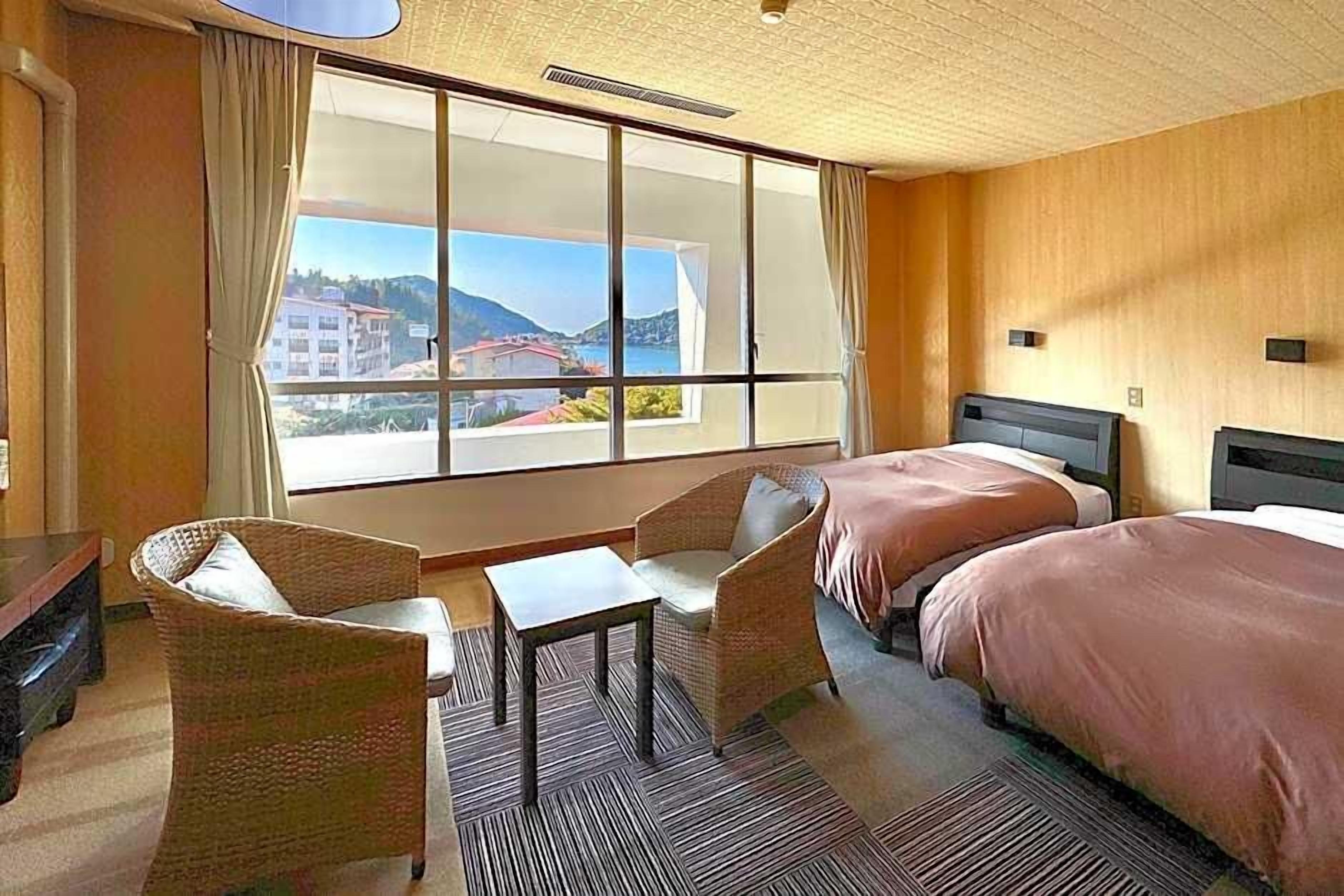 Quarto de hotel com uma janela grande, duas camas, uma mesinha e cadeiras de vime.