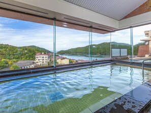 Una piscina interior con paredes de vidrio transparente, vistas a un lago y edificios circundantes.