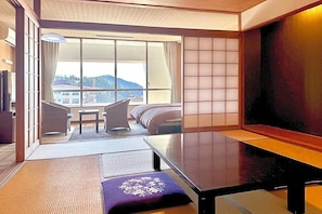 Habitación de hotel con esteras de tatami, una mesa baja, un sofá y un ventanal con vistas a edificios y colinas.