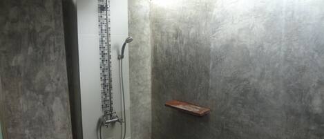 Área de duche com parede revestida em azulejo, chuveiro de metal e prateleira de madeira.