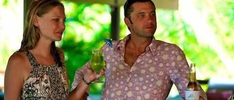 Two people standing at a bar with a green outdoor background.