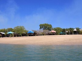 A beachfront property with multiple structures, including a main building and several smaller huts, surrounded by trees and a sandy beach.