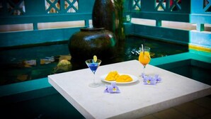 Ein Tisch mit einem blauen Cocktail, einer Mango-Platte und einem Glas Orangensaft, vor einem Hintergrund aus einer Fontäne und einem blauen Zaun.