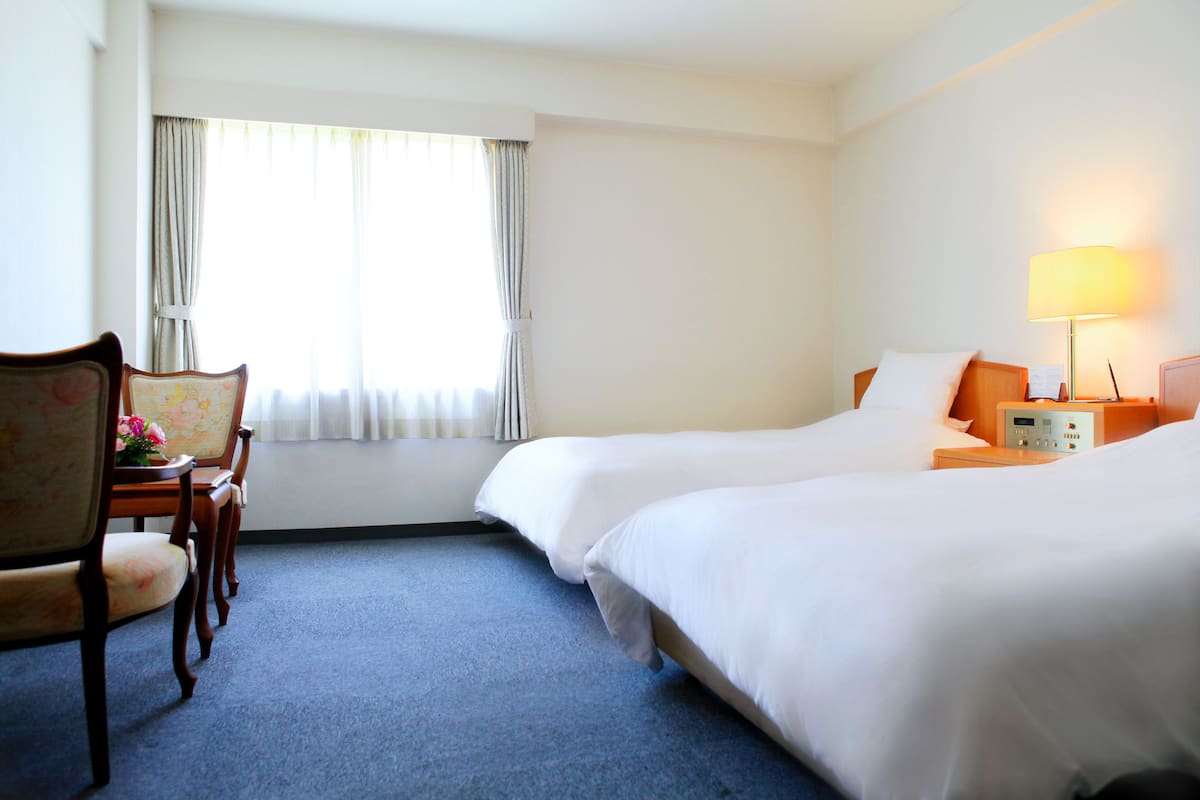 Quarto de hotel com duas camas, uma mesa com cadeira, luminária e janela com cortinas.