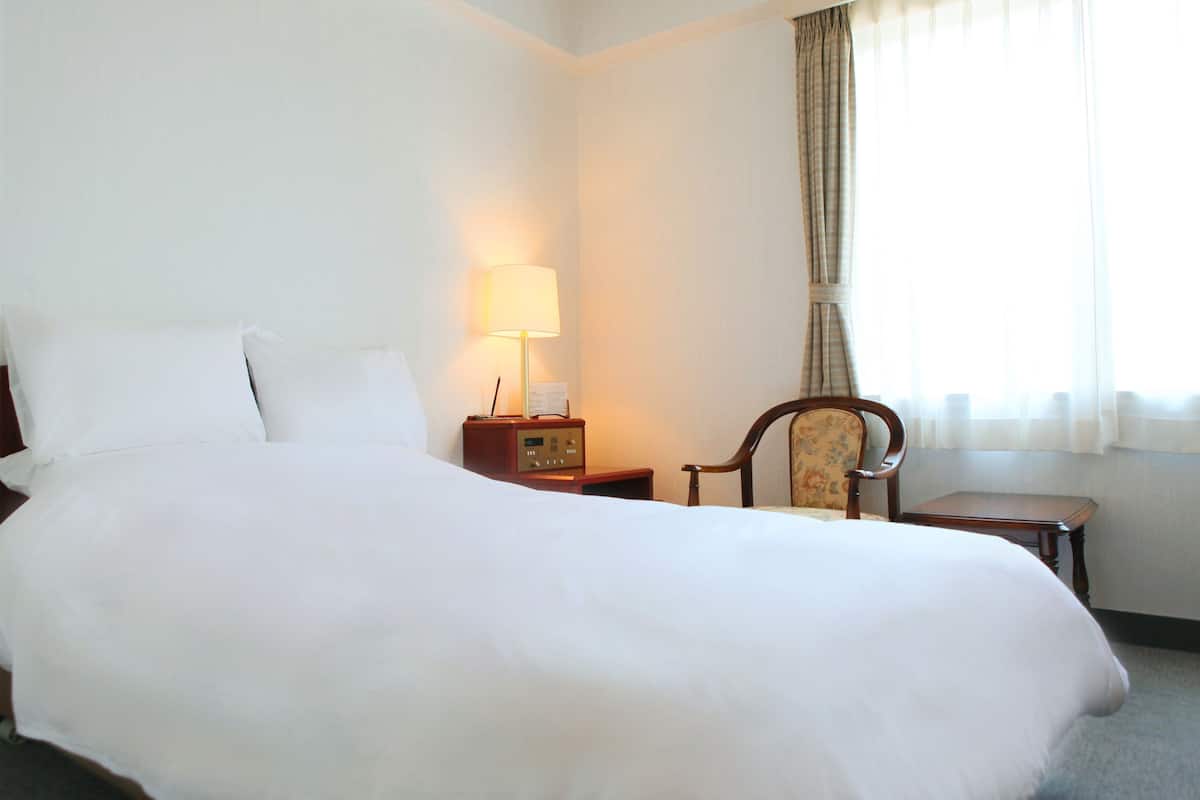Quarto de hotel com uma cama grande, uma mesa de cabeceira com abajur, uma cadeira e uma pequena mesa.