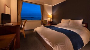 A hotel room with a large bed, a desk, a chair, a lamp, and a view of the ocean at dusk. - Kurashiki Seaside Hotel (Kurashiki)