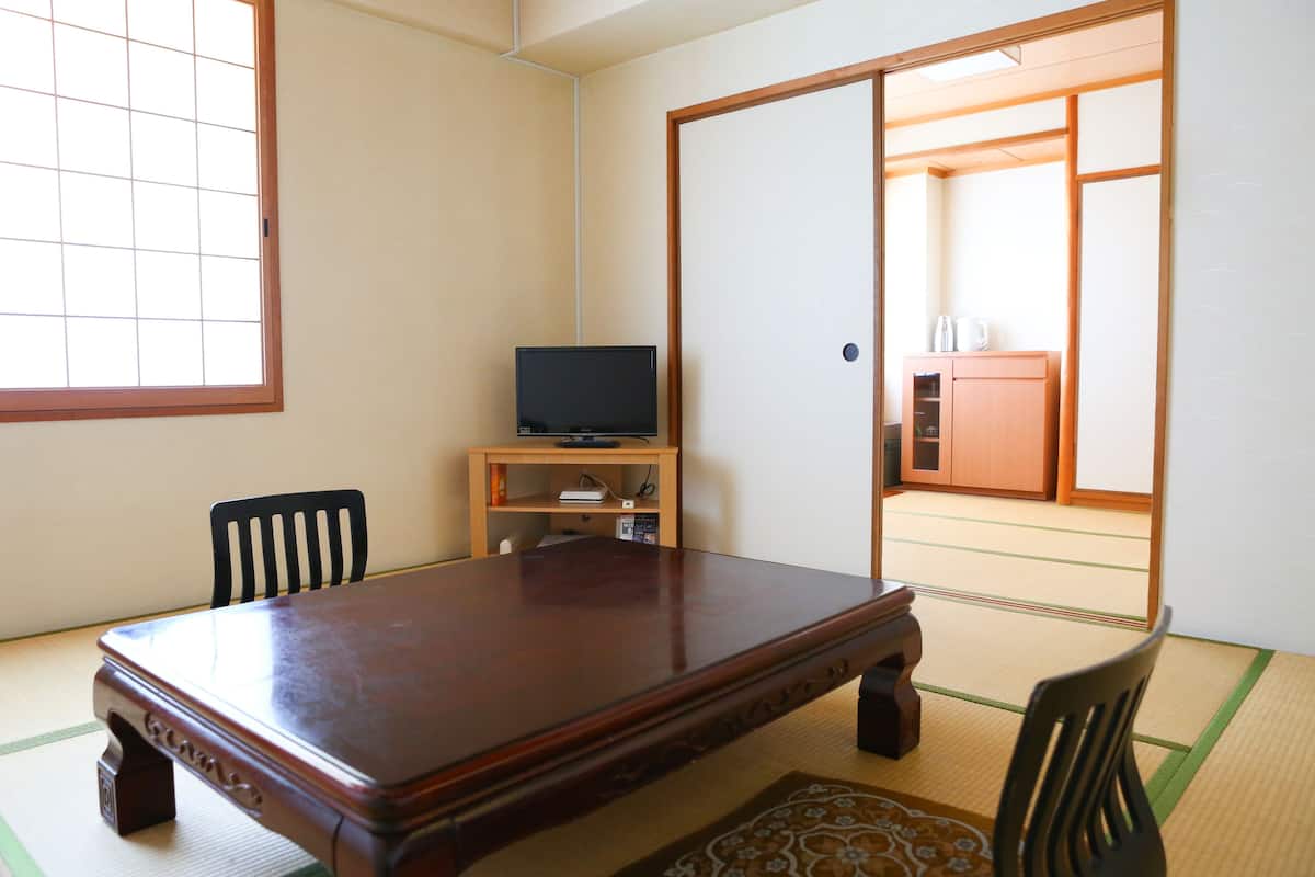 Um quarto japonês tradicional com tatames, uma mesa de madeira, duas cadeiras, uma televisão numa estante e portas deslizantes que dão para uma área de cozinha.