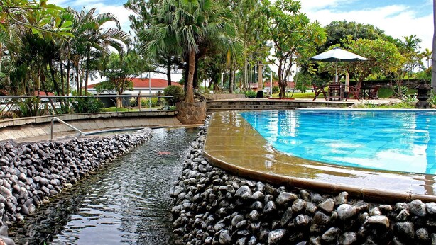 A swimming pool with a stone border, surrounded by tropical plants and a seating area with umbrellas. - Patra Semarang Convention (SEMARANG)