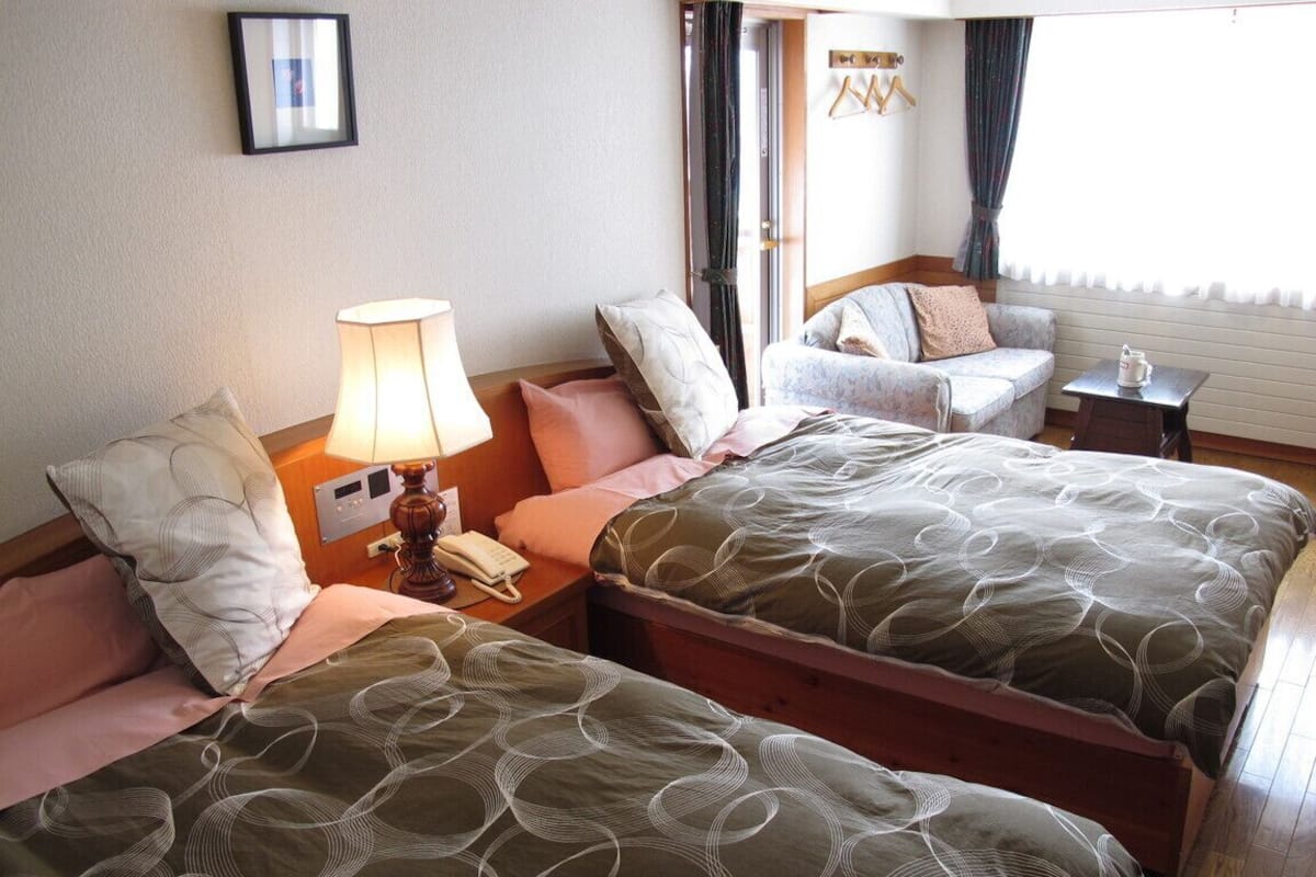 Une chambre d’hôtel avec deux lits, une table de chevet avec une lampe, une chaise et une petite table avec une carafe.