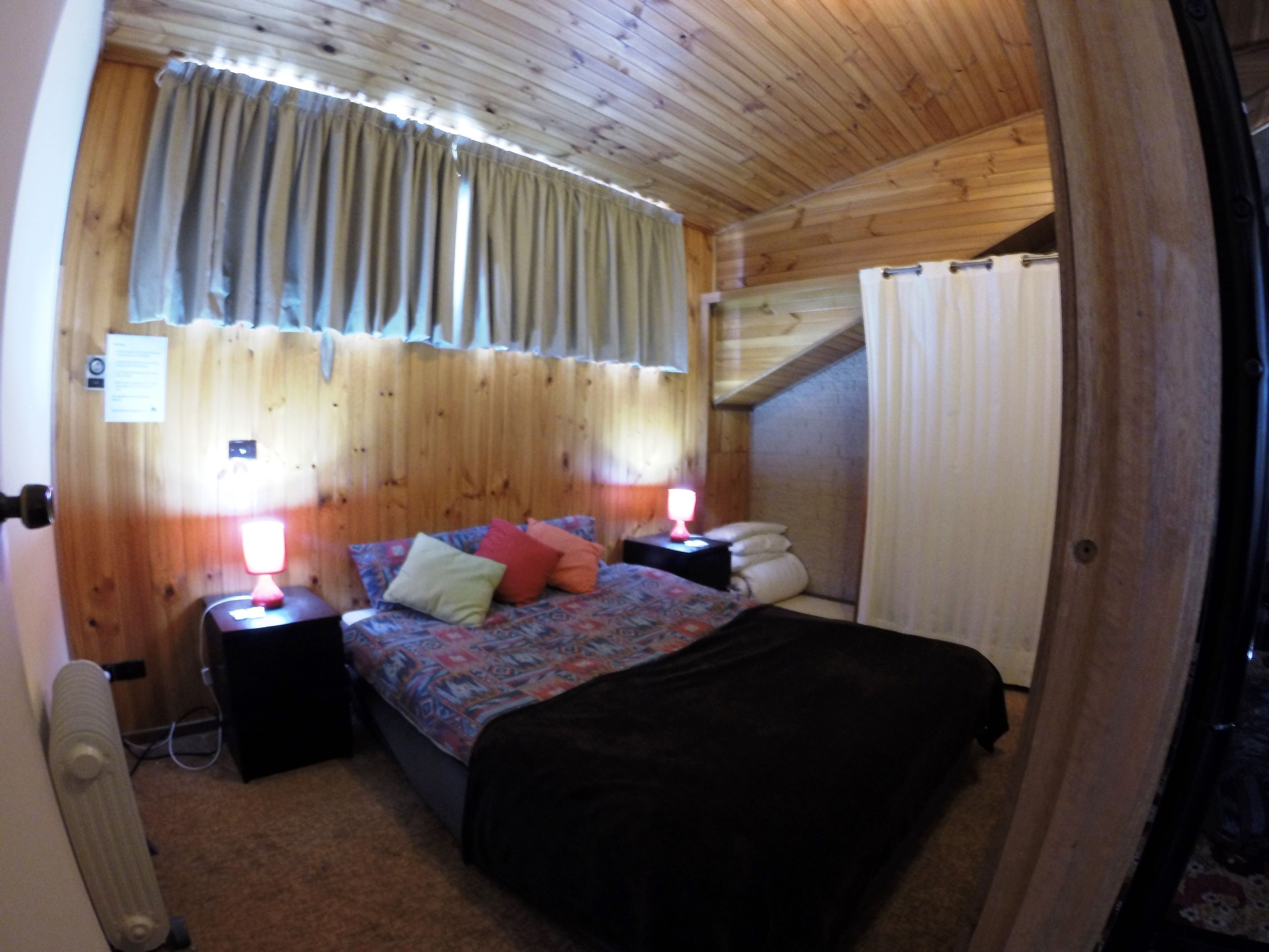 A cozy bedroom with a wooden ceiling, a bed with patterned bedding, two bedside lamps, and a window with curtains.