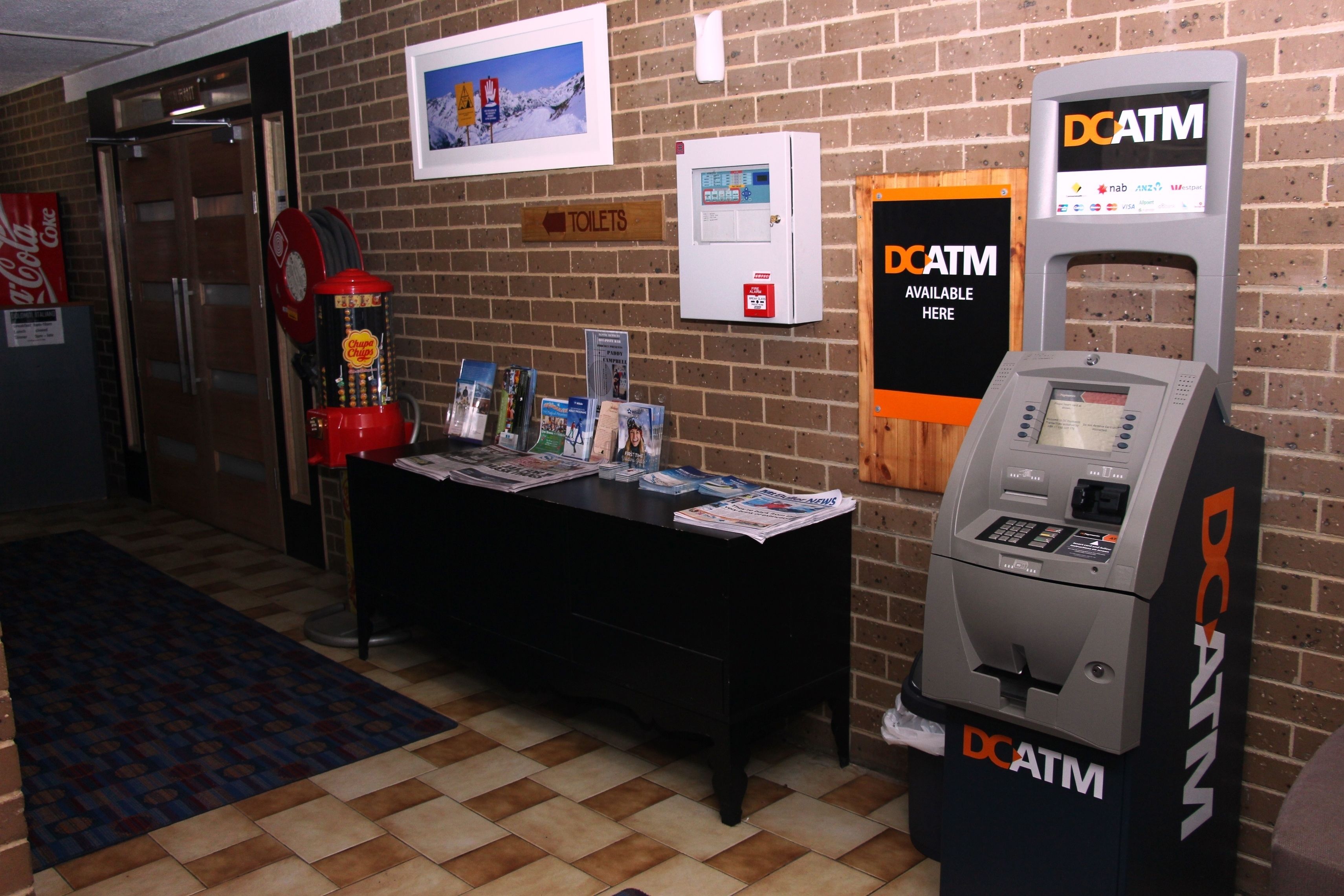 A hotel lobby with a DCATM machine, a sign for toilets, a Coca-Cola refrigerator, and a vending machine.
