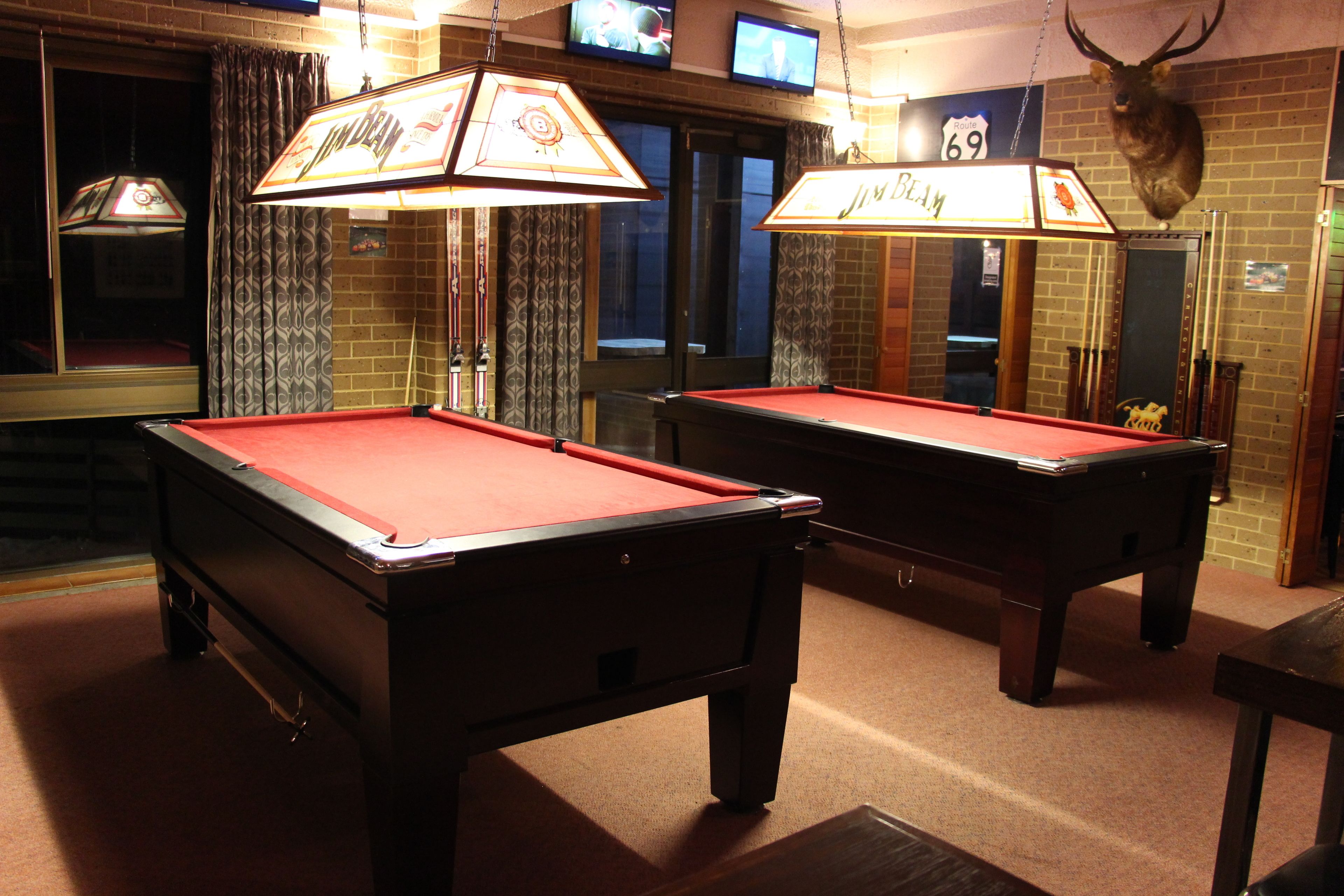 Two pool tables with red felt, overhead lamps, and a mounted deer head on the wall.