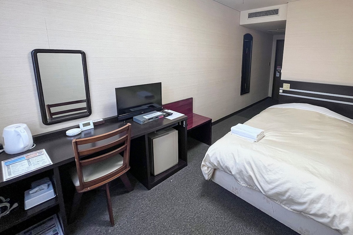 Een hotelkamer met een bed, een bureau met een computer, een stoel, een spiegel en een kleine koelkast.