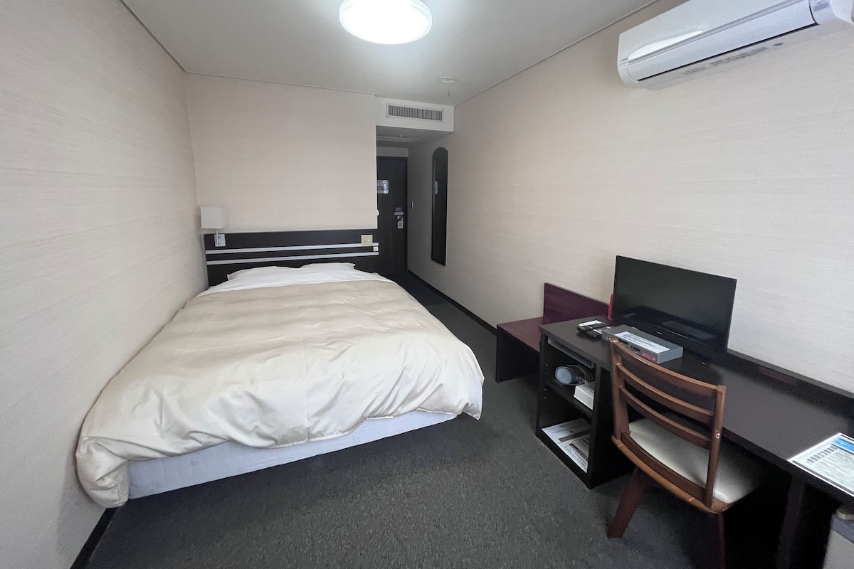 Een kleine hotelkamer met een bed, een bureau met stoel, een televisie en een airconditioner.