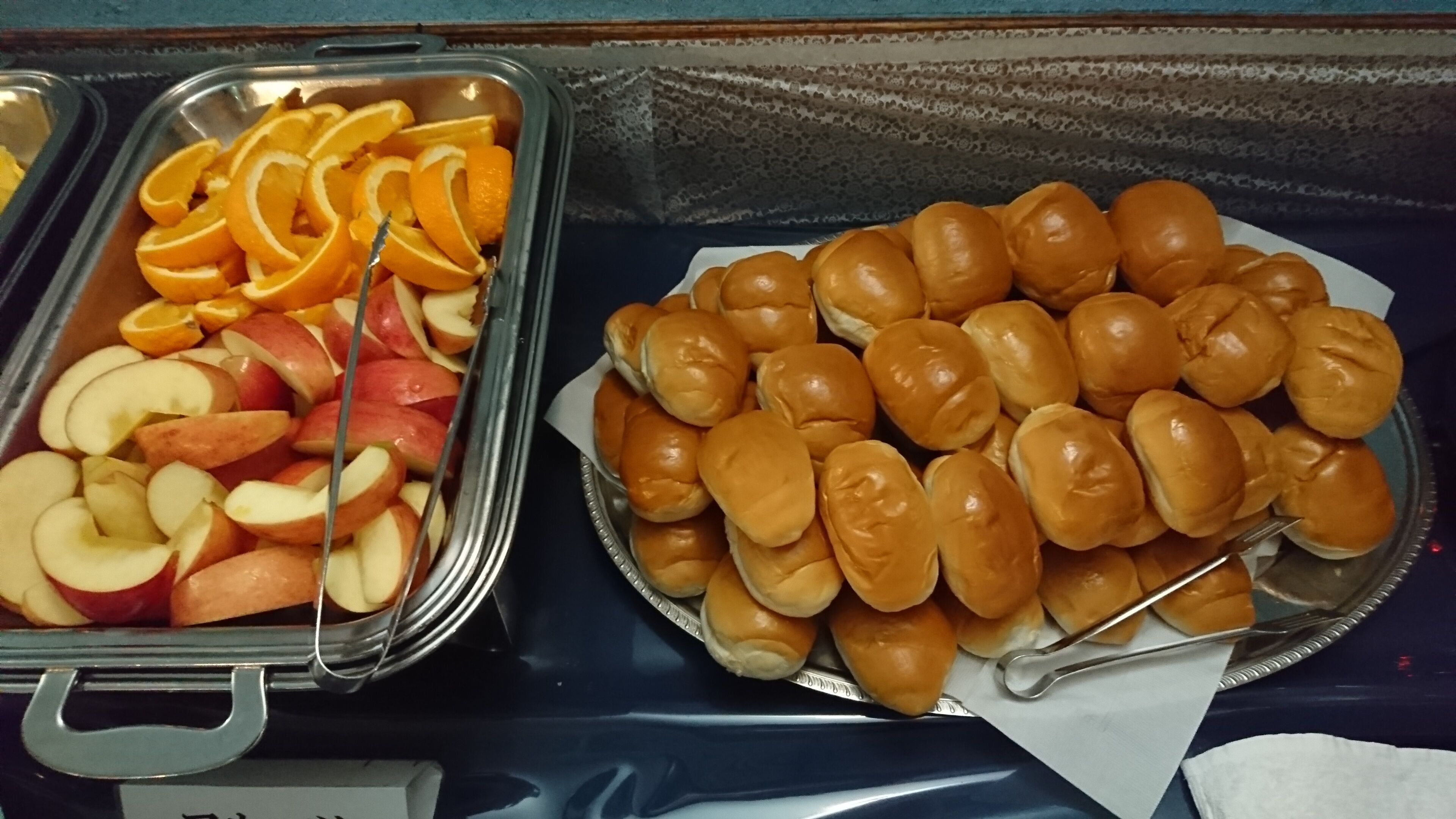 A buffet with trays of sliced apples and oranges, and a tray of rolls.