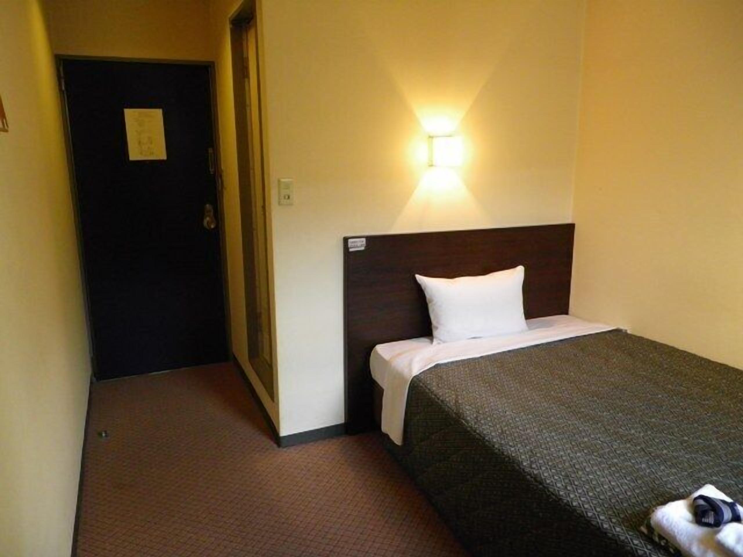 Habitación de hotel con una cama individual, una mesita de noche, una lámpara fijada a la pared y una puerta que da a otra habitación.