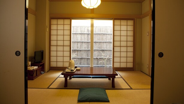 Una habitación tradicional japonesa con suelo de tatami, una mesa baja, un televisor y puertas correderas que dan a una valla de bambú.