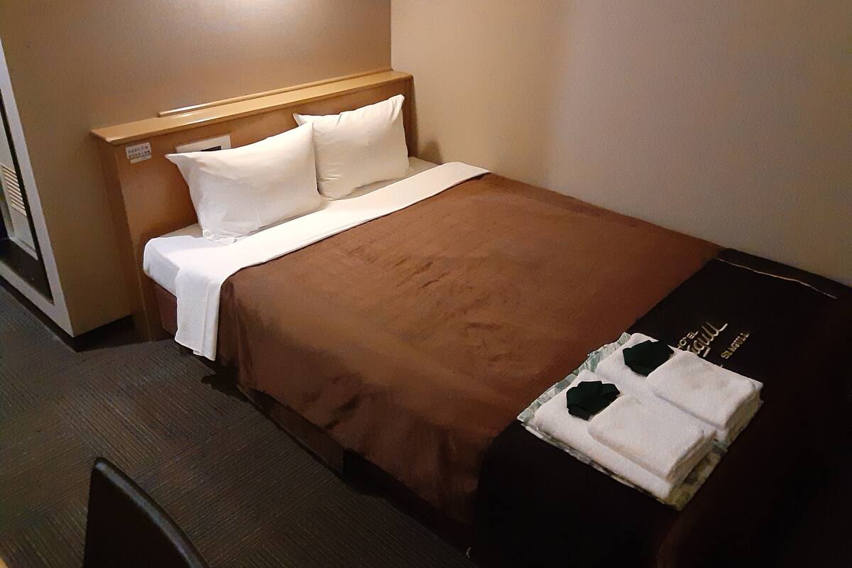 A hotel room with a bed, a wooden headboard, a brown bedspread, white pillows, and a black suitcase on the floor.