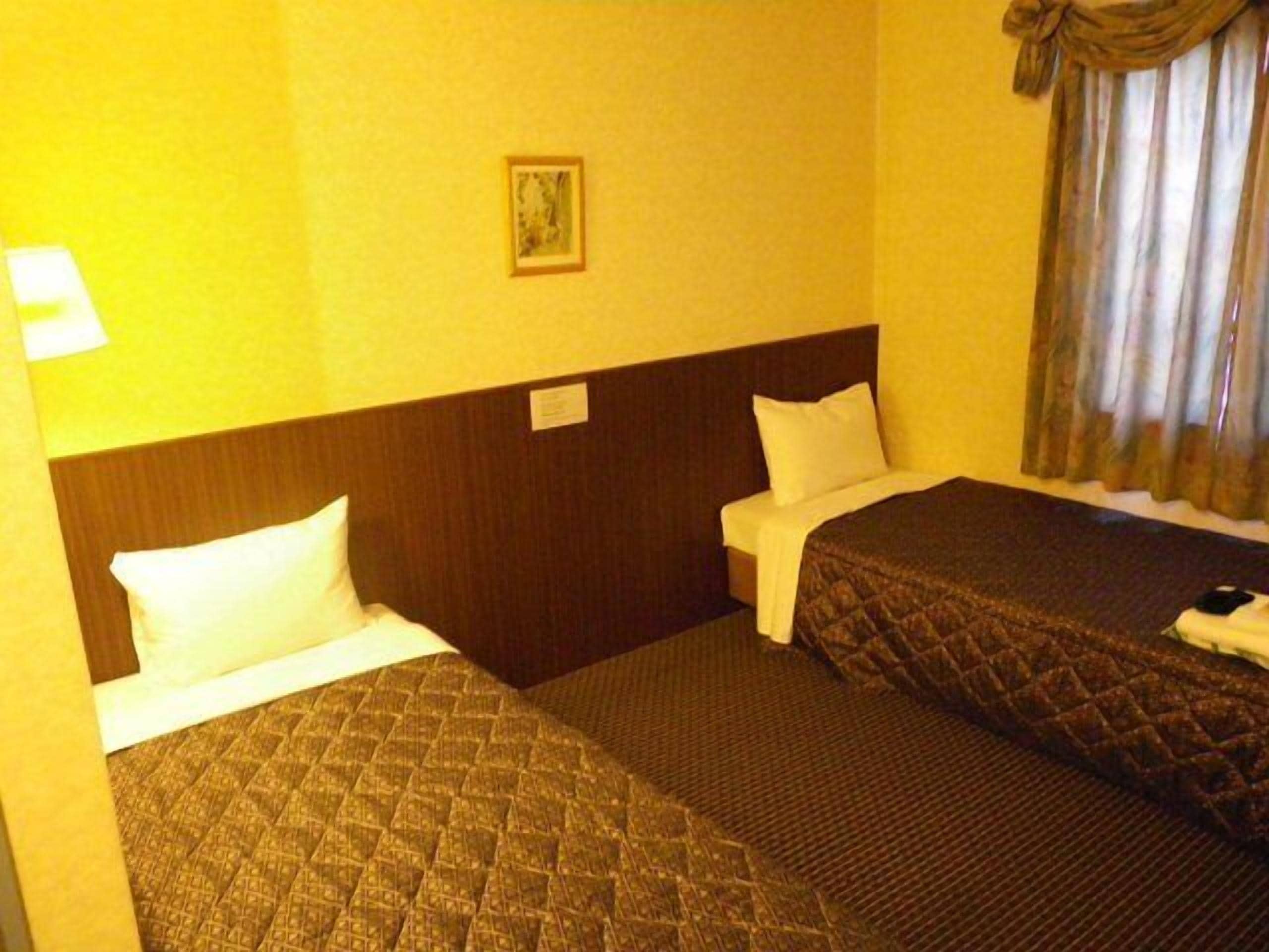 Una habitación de hotel con dos camas, cabeceros, mesitas de noche, una lámpara, un cuadro enmarco y una ventana con cortinas.