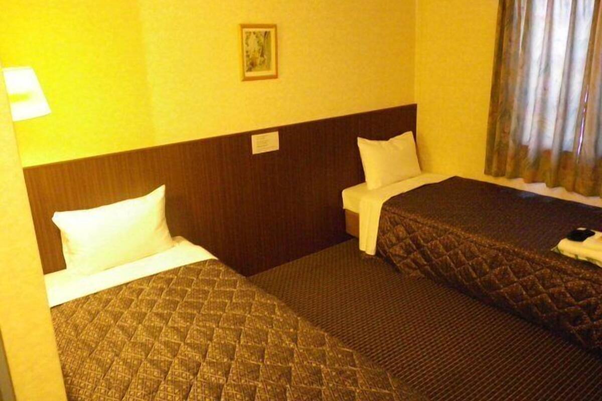 A hotel room with two beds, a headboard, a nightstand, a lamp, a framed picture, and a window with curtains.