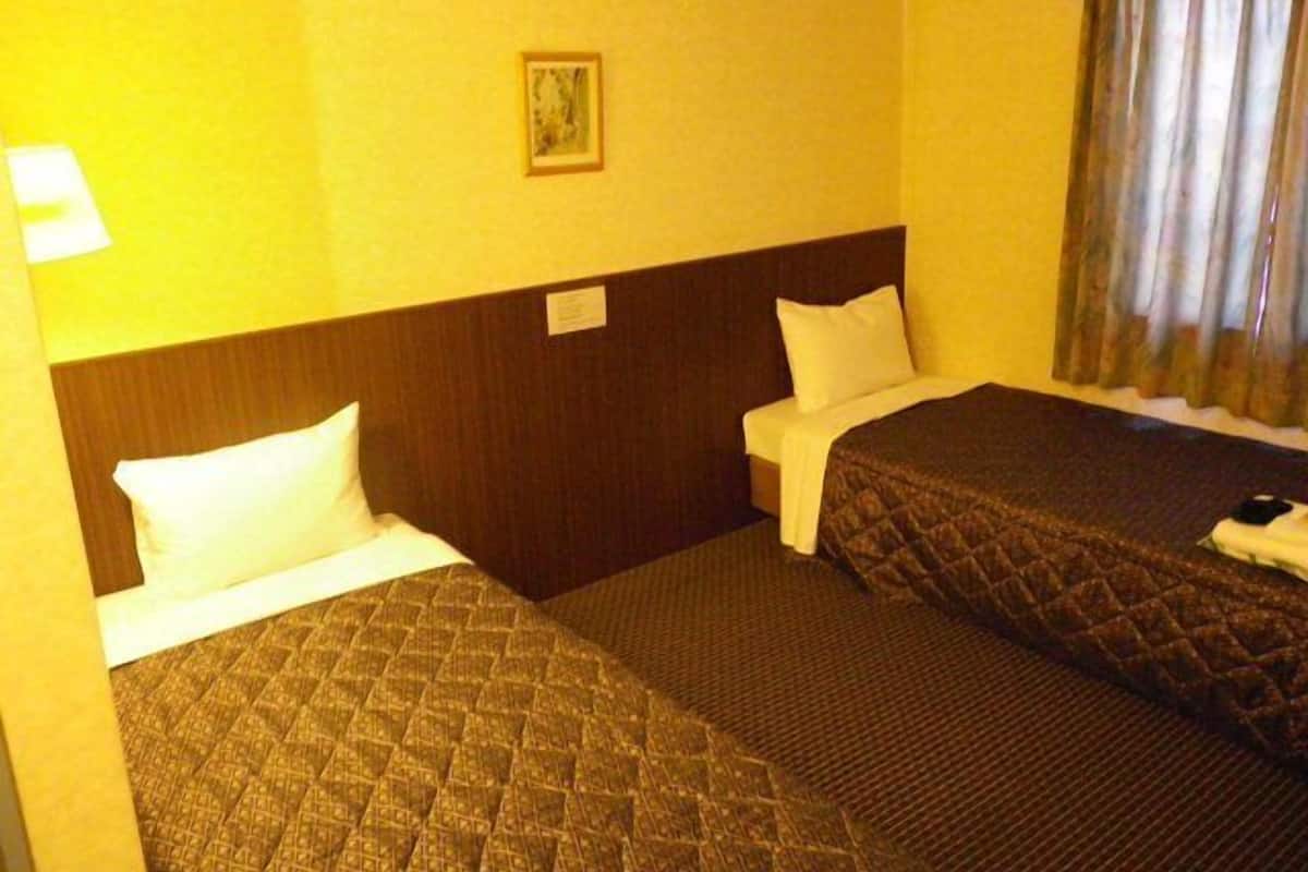 Una habitación de hotel con dos camas, cabeceros, mesitas de noche, una lámpara, un cuadro enmarco y una ventana con cortinas.