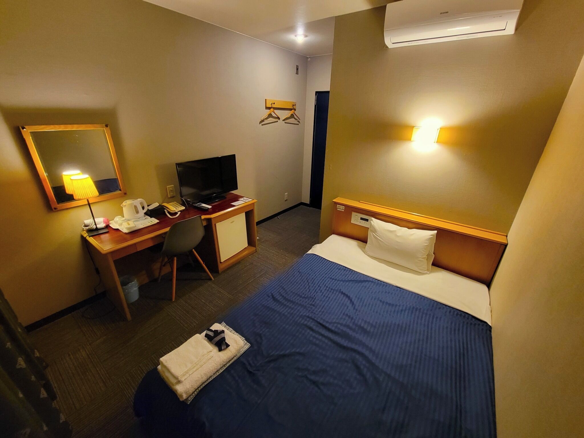 Habitación de hotel con cama, escritorio con televisor, una silla, lámpara de escritorio y equipo de aire acondicionado.