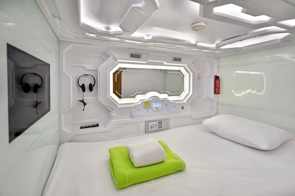 A modern, white-themed room with a bed, a green towel, and a pair of headphones on the wall.