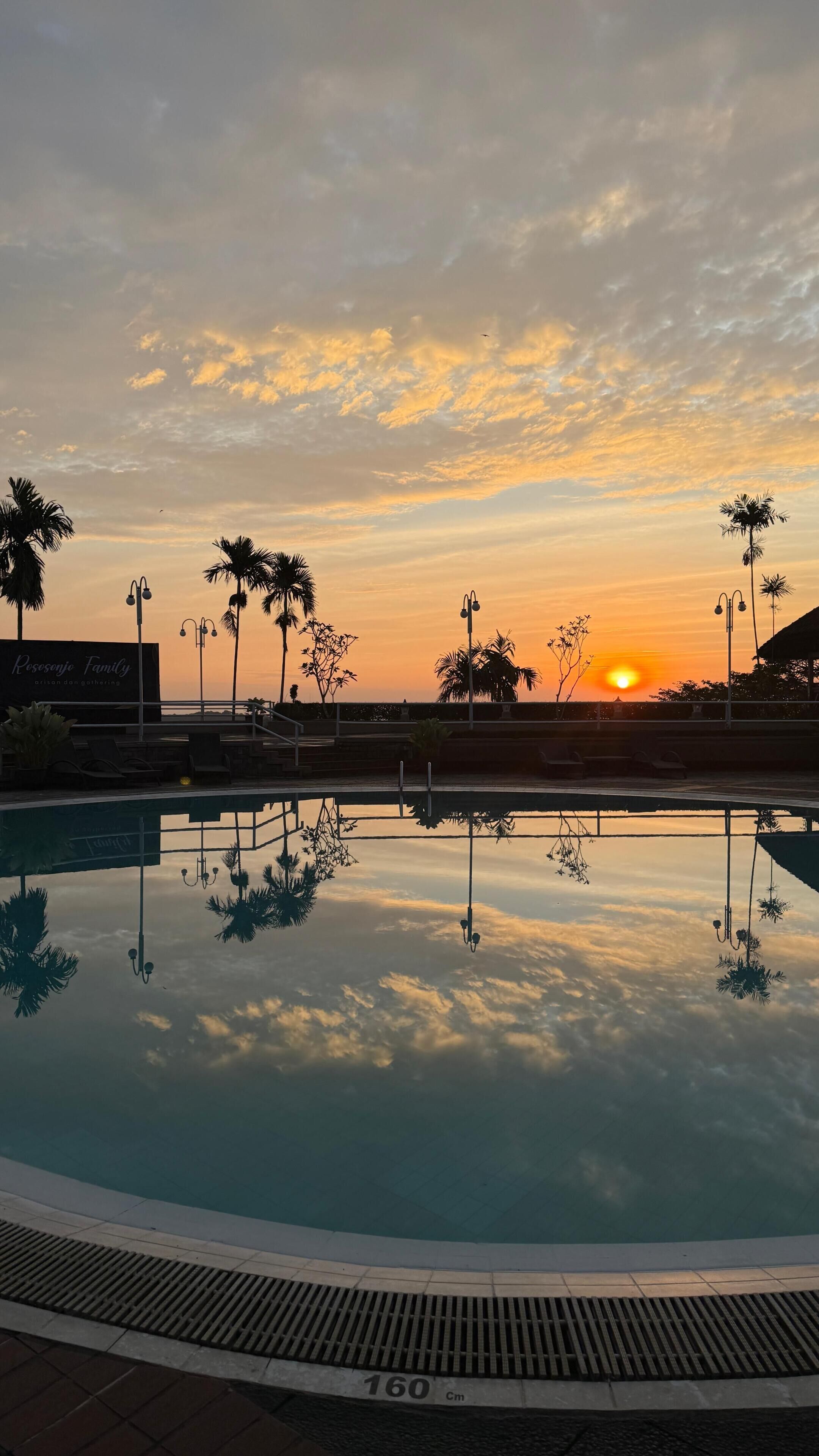 Piscina refletindo o pôr do sol, palmeiras e uma placa com 'Rosesje Family'.