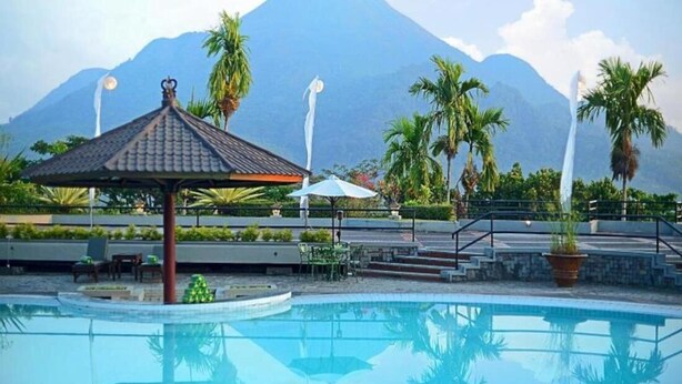 A swimming pool with a gazebo, palm trees, and a mountain in the background. - Grand Whiz Hotel Trawas (Trawas)