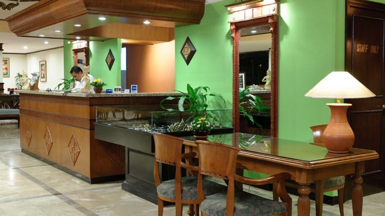 A hotel reception area with a wooden reception desk, a glass-top table, and a chair. There is a large mirror above the desk and a green wall with a 'STAFF ONLY' sign.