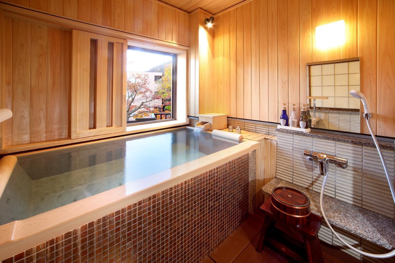 A hot tub with a view of a tree outside, wooden walls, a tiled shower area, and a small wooden stool.