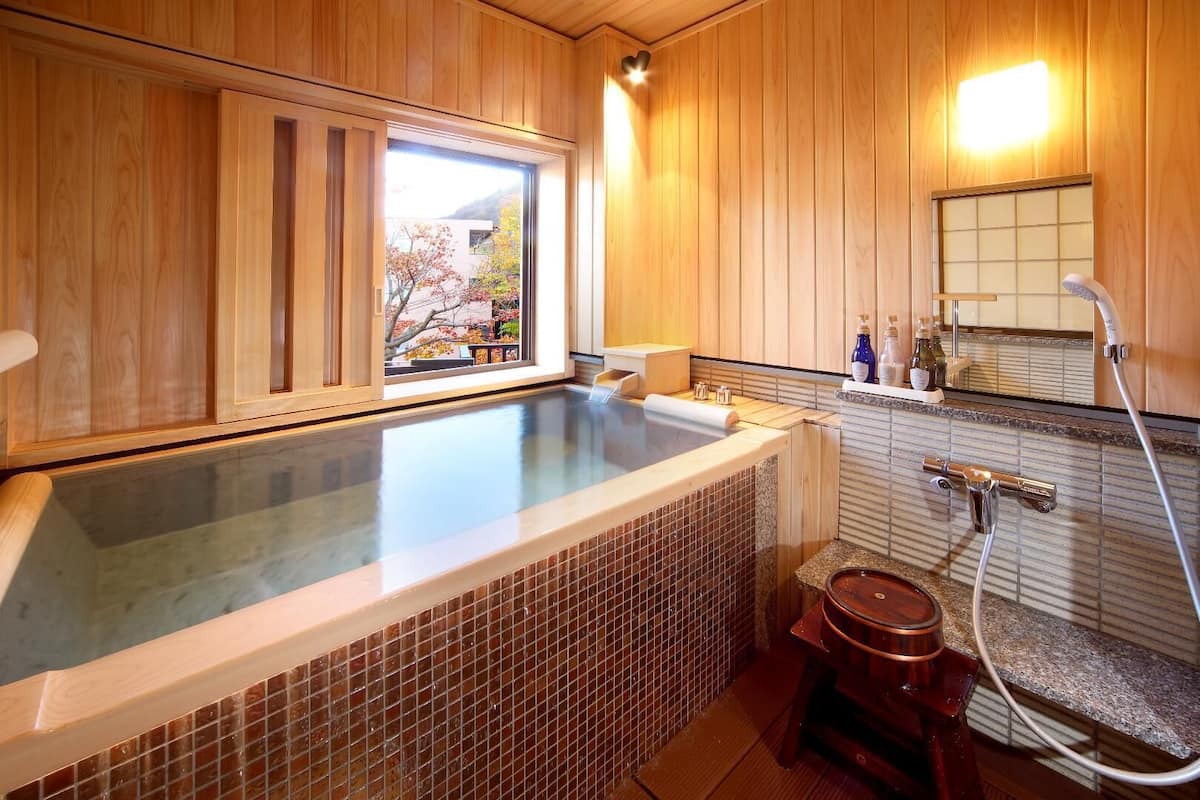 Un jacuzzi con vista a un árbol, paredes de madera, zona de ducha con azulejos y un pequeño taburete de madera.