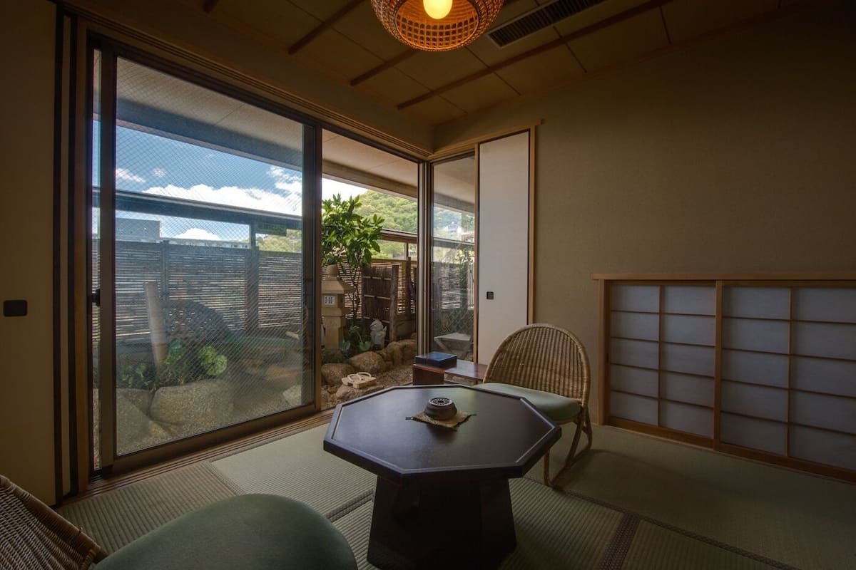 Una habitación con suelo de tatami, una mesa baja con un objeto decorativo, una silla, una puerta corredera con vistas al exterior y un biombó japonés tradicional.
