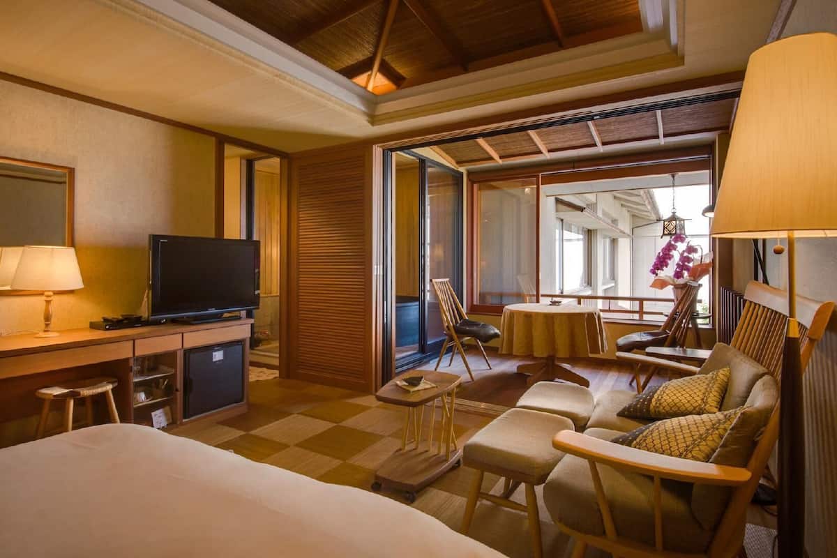 Habitación de hotel con cama, escritorio con televisor, zona de comedor con sillas y vista por un ventanal grande.