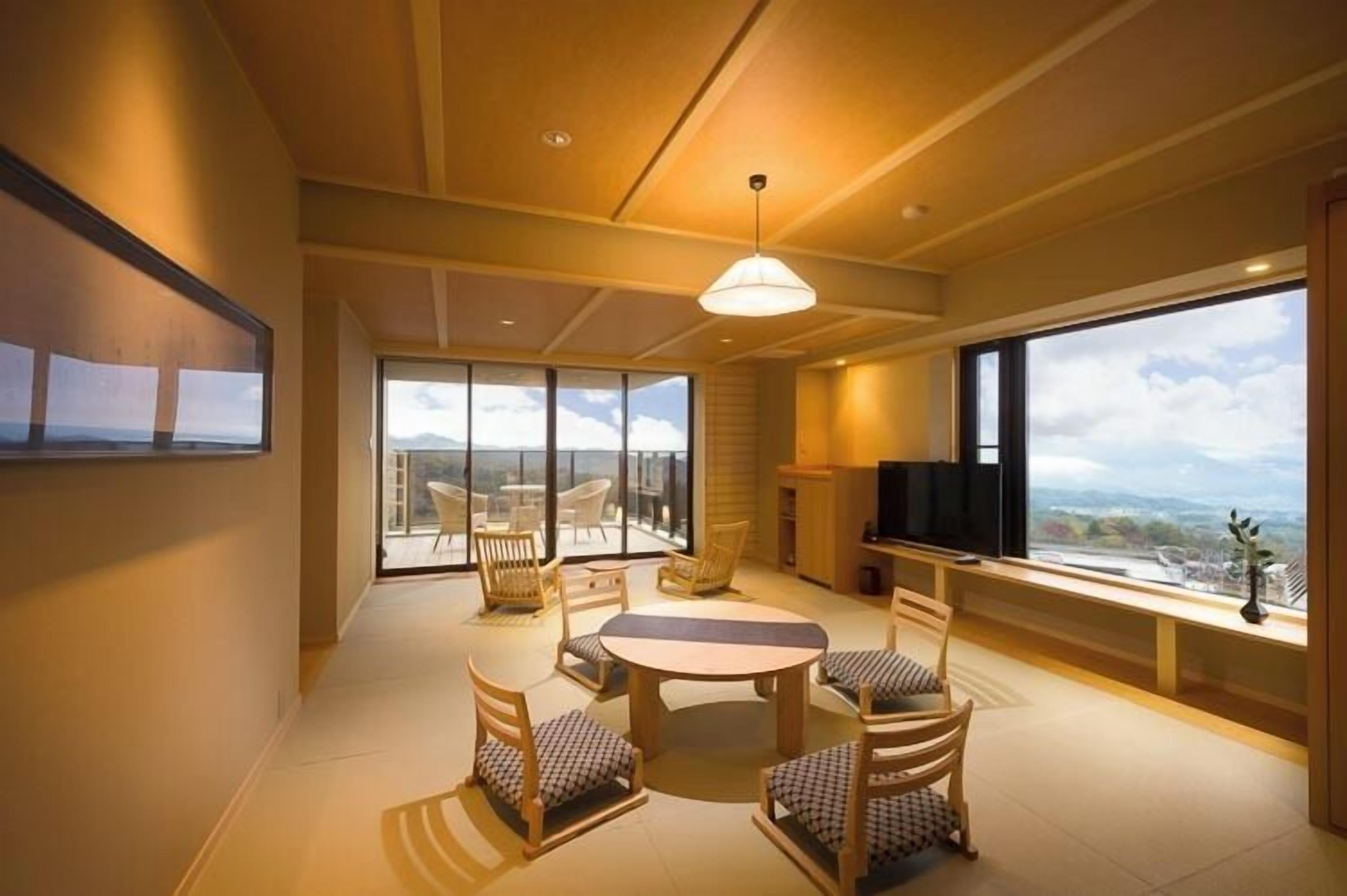 A modern living room with a round table and chairs, a TV, and large windows offering a view of the mountains.