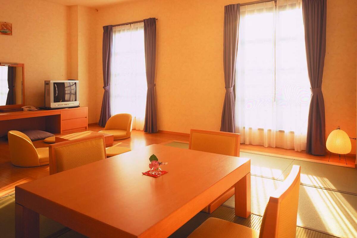 A hotel room with a wooden dining table, two chairs, a sofa, a TV, and large windows with curtains.