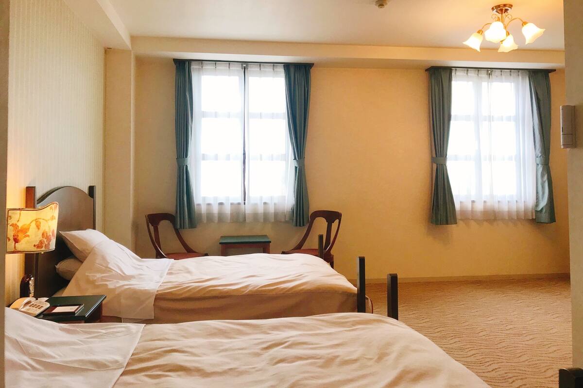A hotel room with two beds, a nightstand with a lamp, two chairs, and a window with curtains.