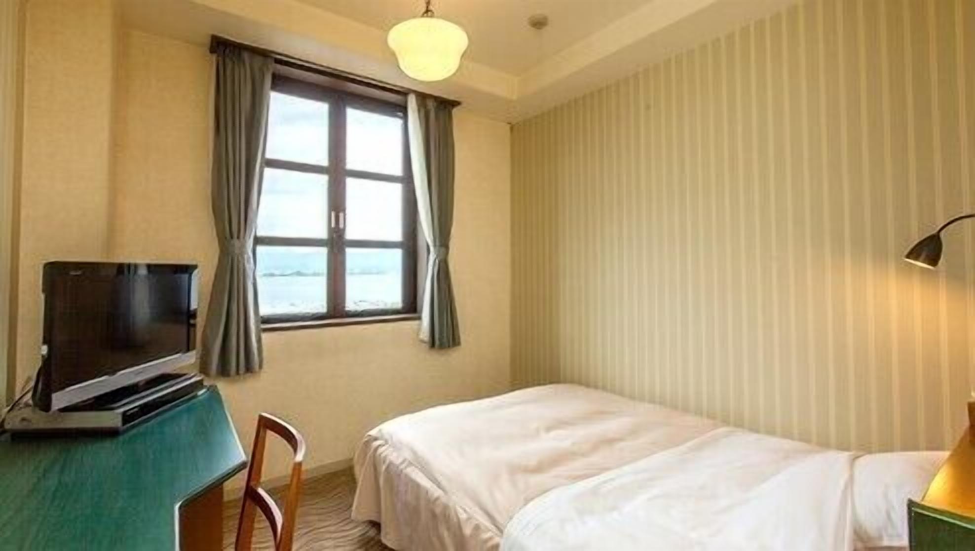 A hotel room with a bed, a desk with a television, a chair, a window with curtains, and a lamp.