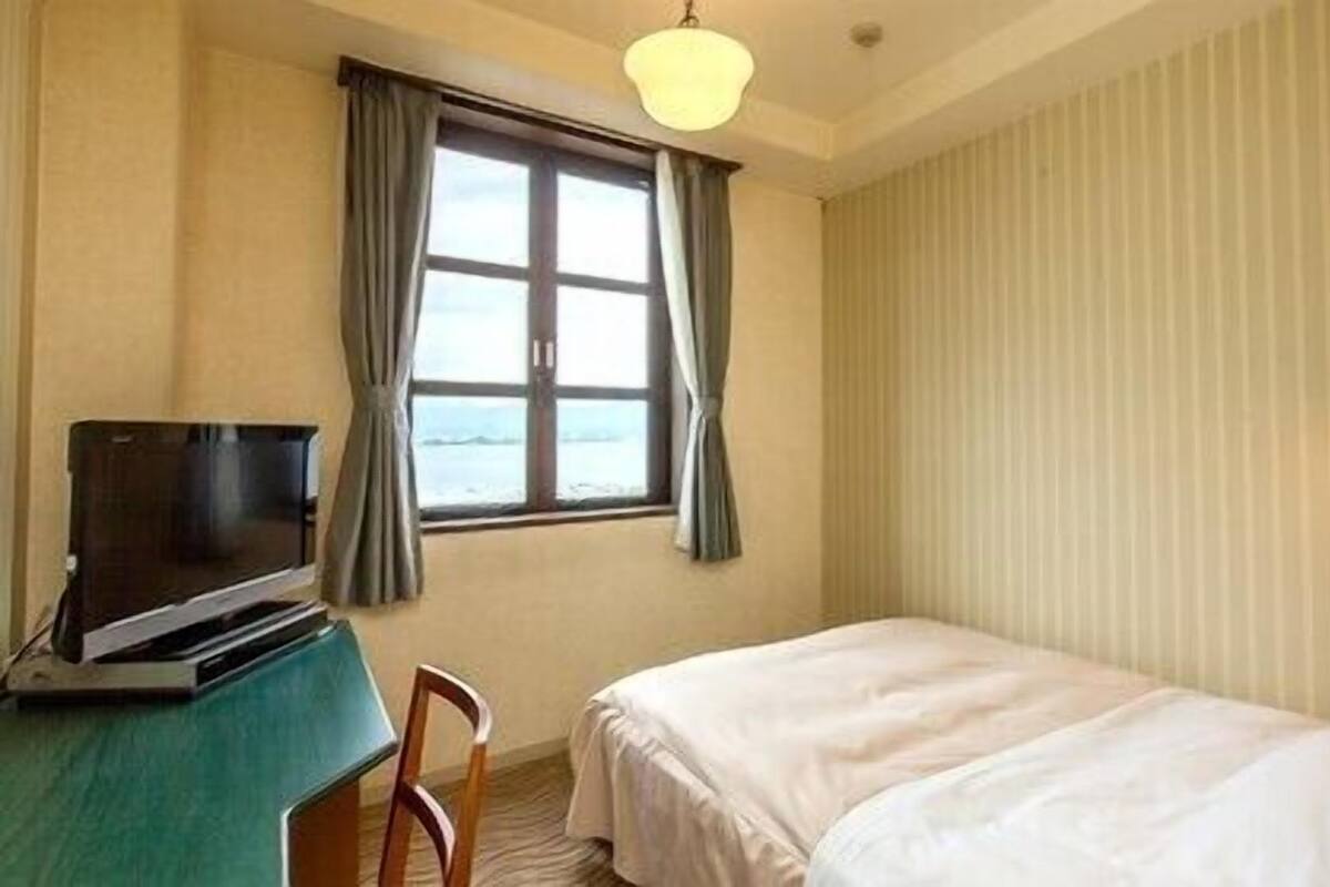 A hotel room with a bed, a desk with a television, a chair, a window with curtains, and a lamp.