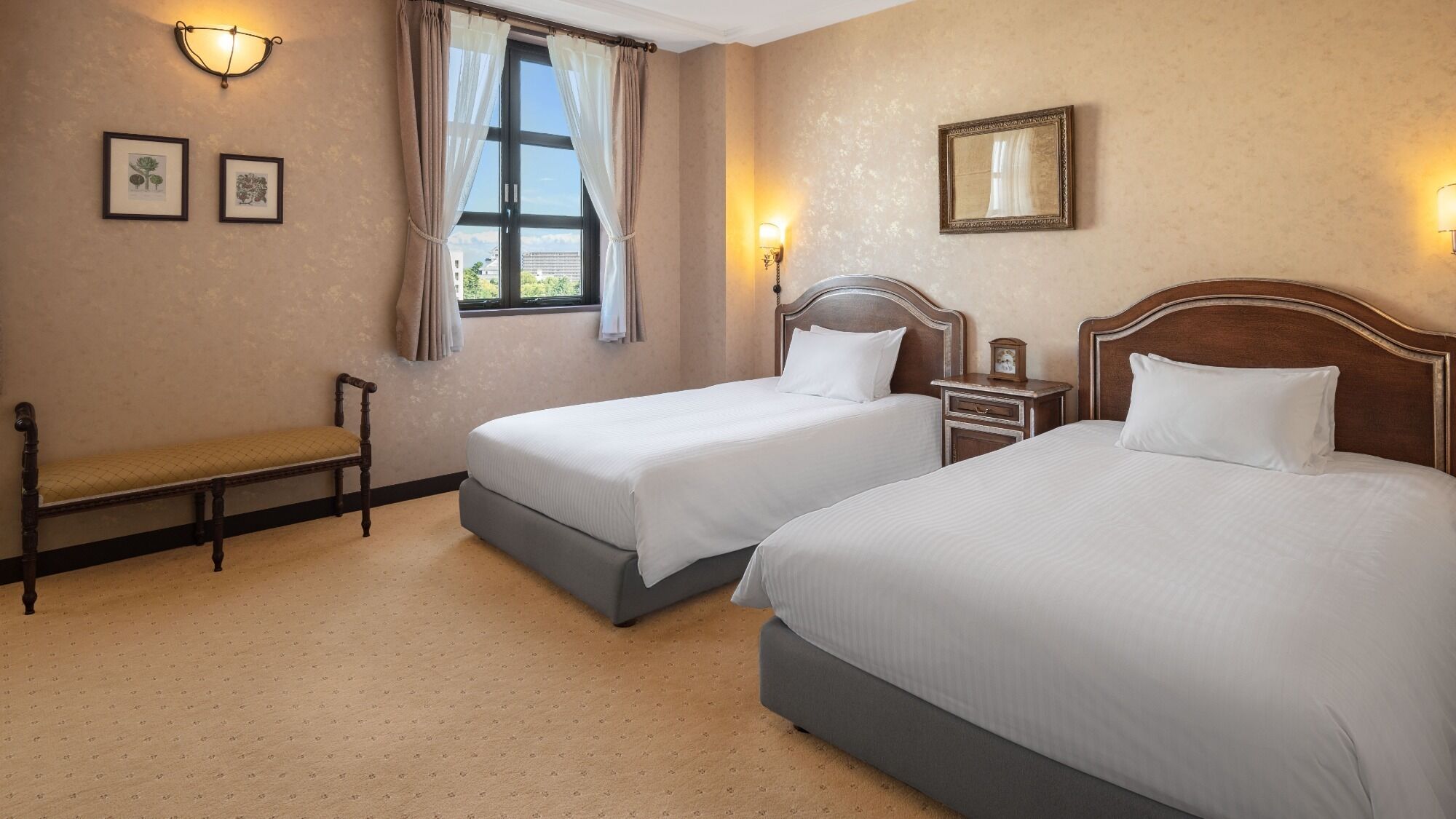 A hotel room with two beds, a bench, a window with curtains, wall-mounted lights, and framed botanical prints.