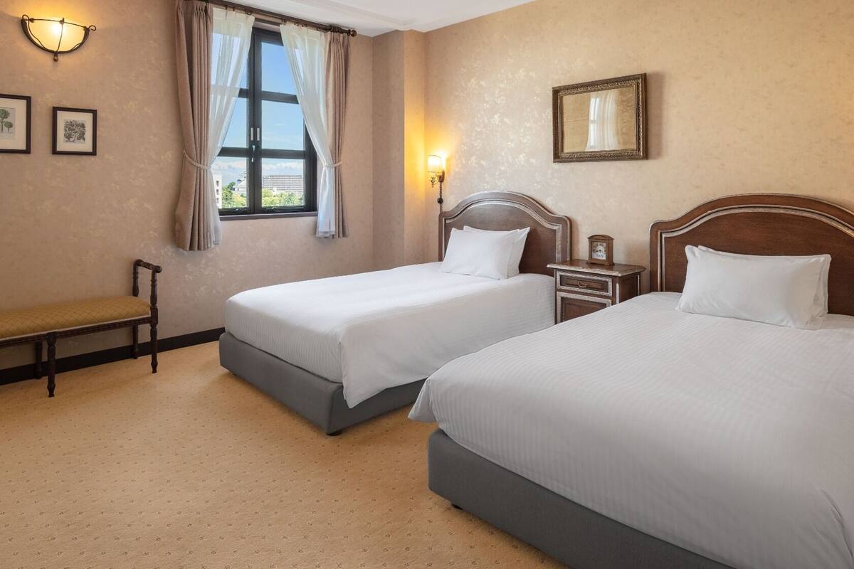 A hotel room with two beds, a bench, a window with curtains, wall-mounted lights, and framed botanical prints.