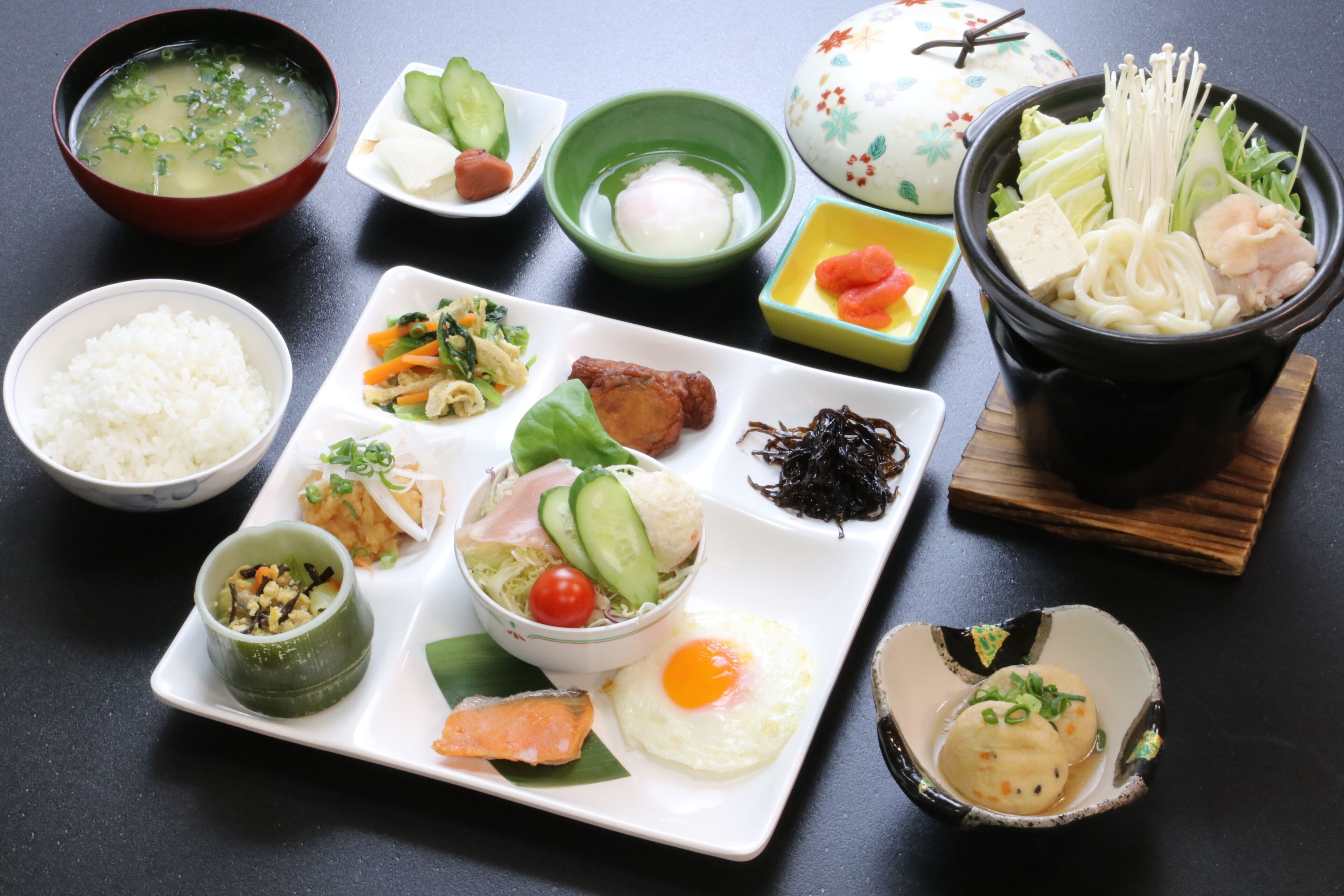 Verschiedene japanische Gerichte, darunter Reis, Gemüse und Meeresfrüchte, auf einem schwarzen Tisch angerichtet.