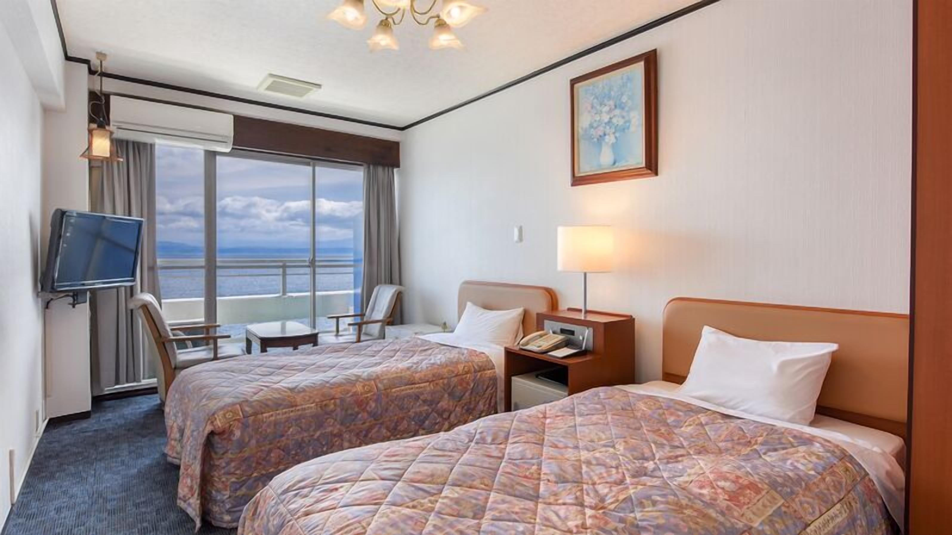 A hotel room with two beds, a TV, a desk, and a view of the sea.