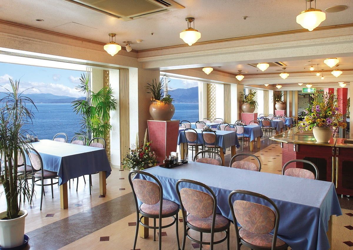 Un restaurante con mesas y sillas, amplias ventanas con vista al mar y plantas en macetas.