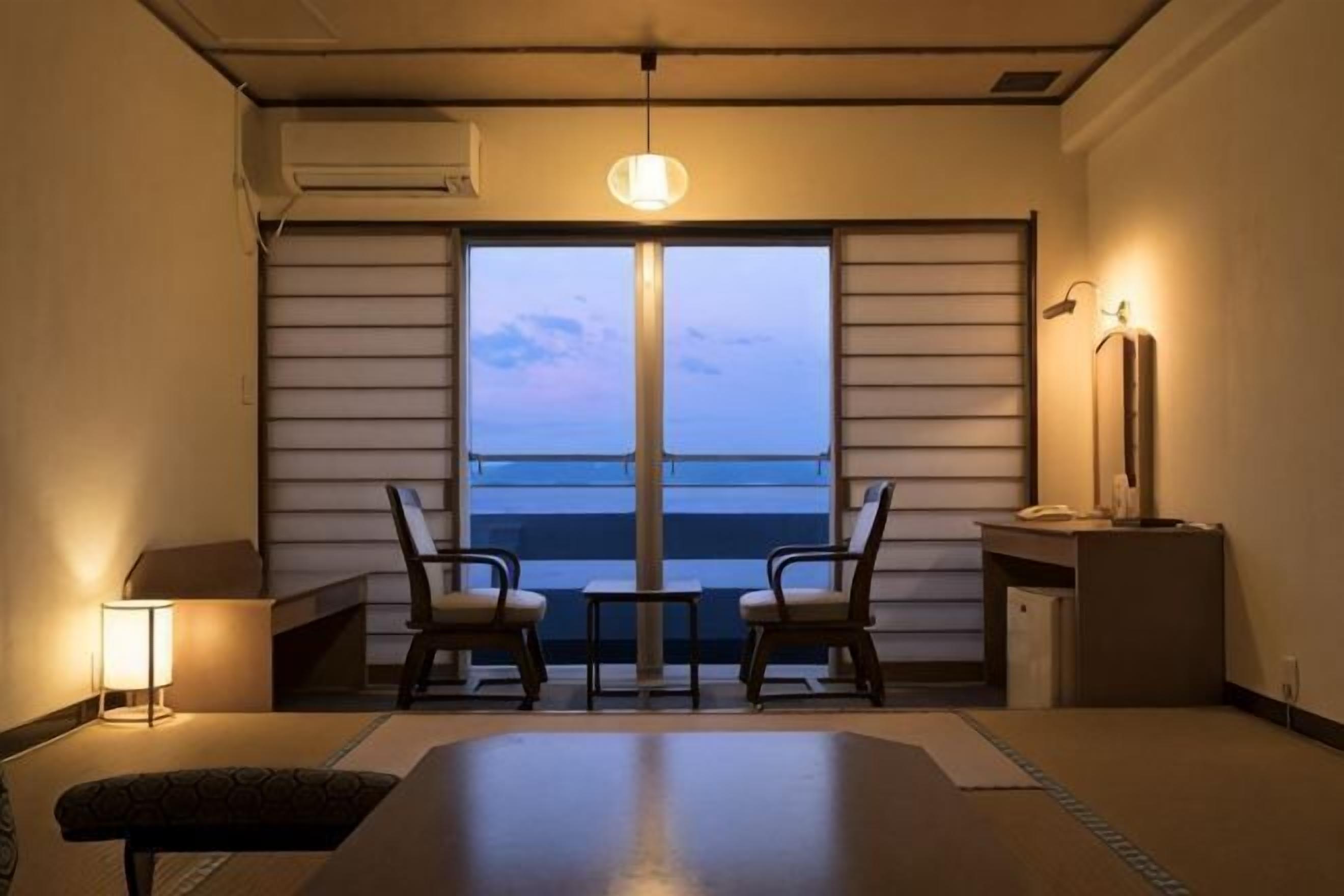 Una habitación con un ventanal grande, dos sillas, una mesa, una lámpara y un escritorio con lámpara.