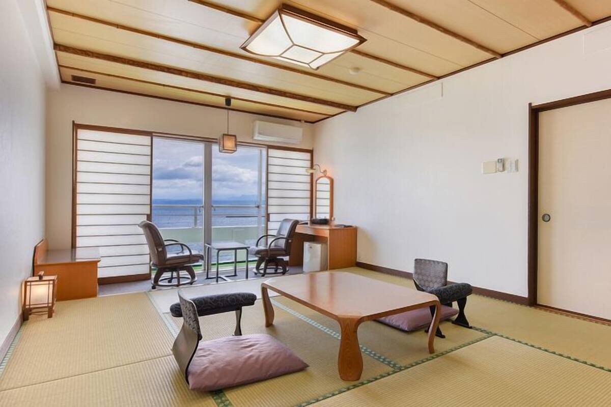 Una habitación con suelo de tatami, una mesa baja, sillas, vistas a través de amplios ventanales y una puerta corredera.