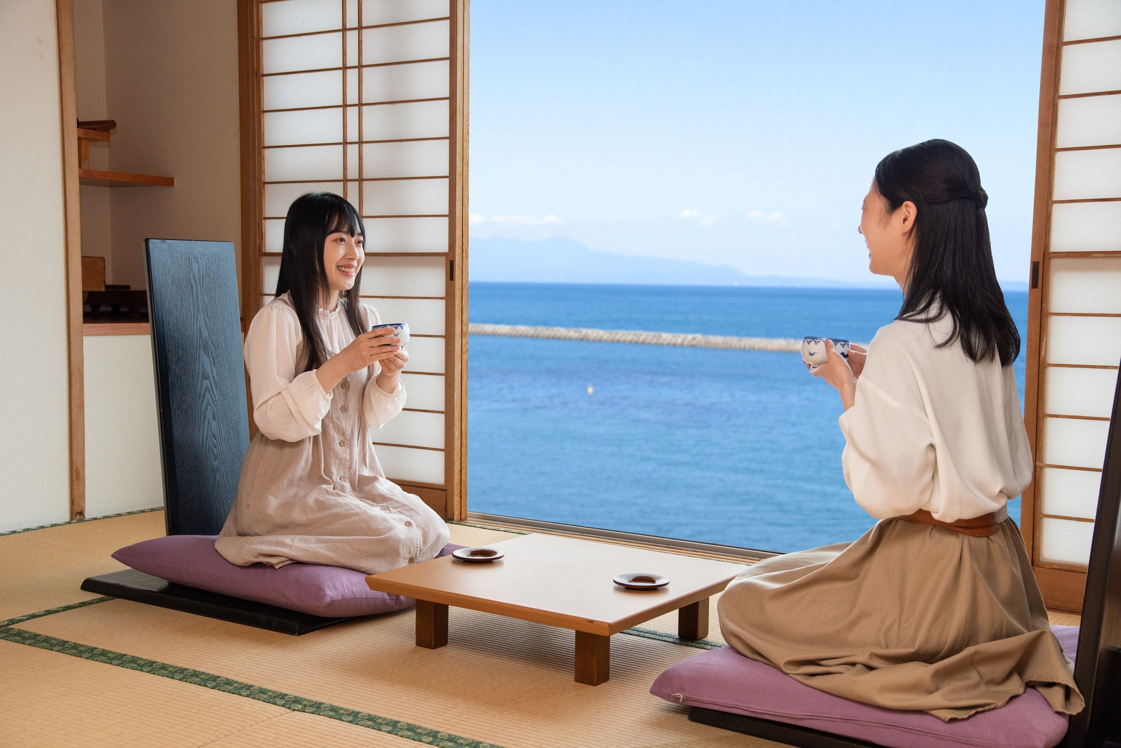Two people sitting on tatami mats facing each other with a view of the sea through a shoji screen.