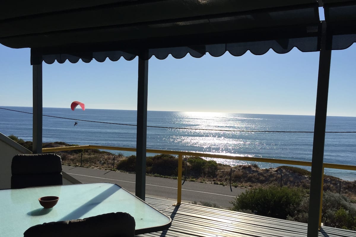 A covered outdoor seating area with a view of the ocean and a paraglider in the distance.