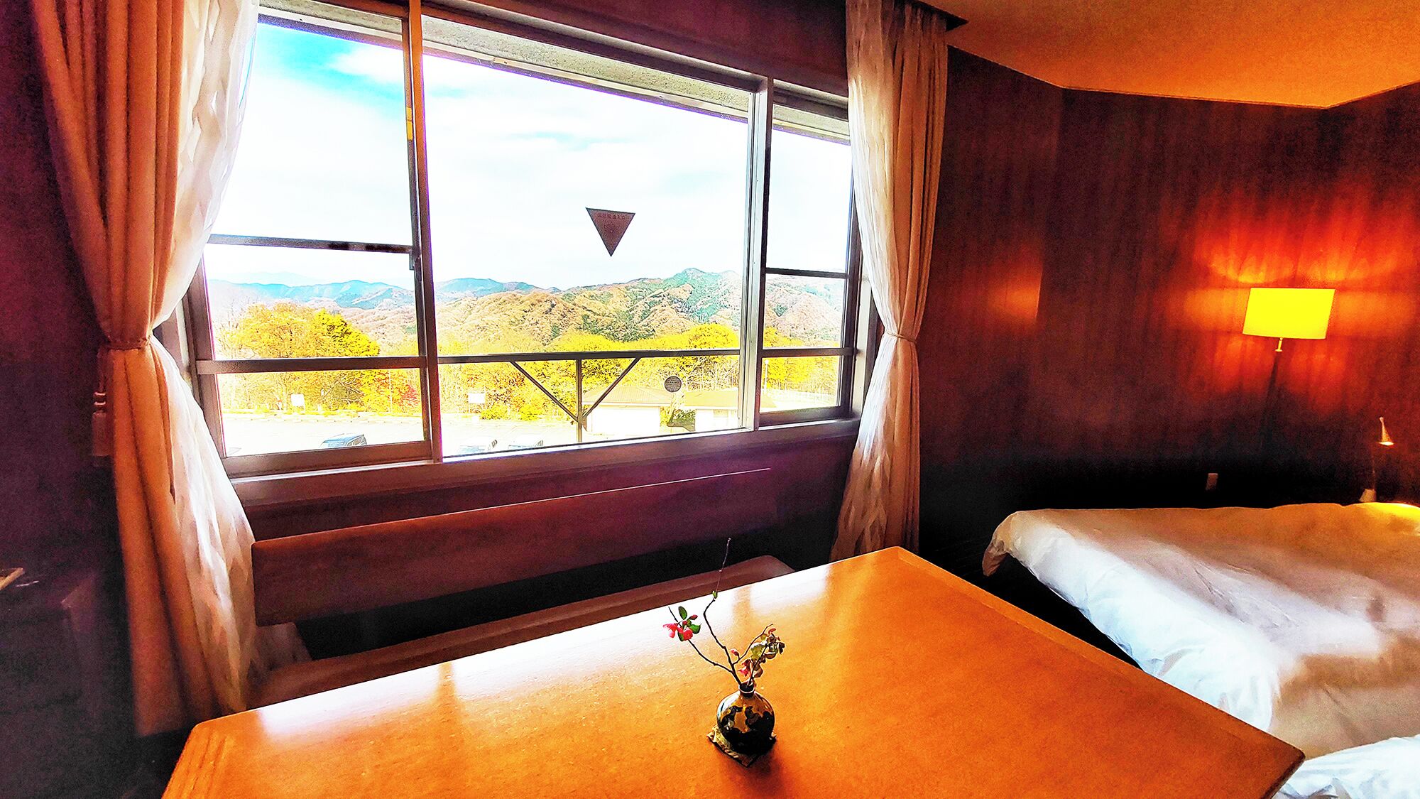 A hotel room with a bed, a small table with a vase, a window with curtains, and a view of a mountainous landscape.